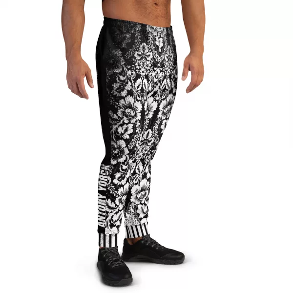 Ein Mann steht vor einem weißen Hintergrund und trägt eine schwarze Jogginghose der Marke ANTONY YORCK mit einem auffälligen weißen floralen Muster im baroque Noir Stil. Die Hose weist den Schriftzug "ANTONY YORCK" am Bein auf und hat ein gestreiftes Bündchen. Der Mann kombiniert sie mit schwarzen Sportschuhen. Perfekt für luxury urban street style, streetwear fashion, subversive style und hiphop style Liebhaber. Entdecken Sie mehr auf https://antonyyorck.com. Schlüsselwörter: trendy menswear, modern fashion, designer joggers, urban clothing, stylish activewear.