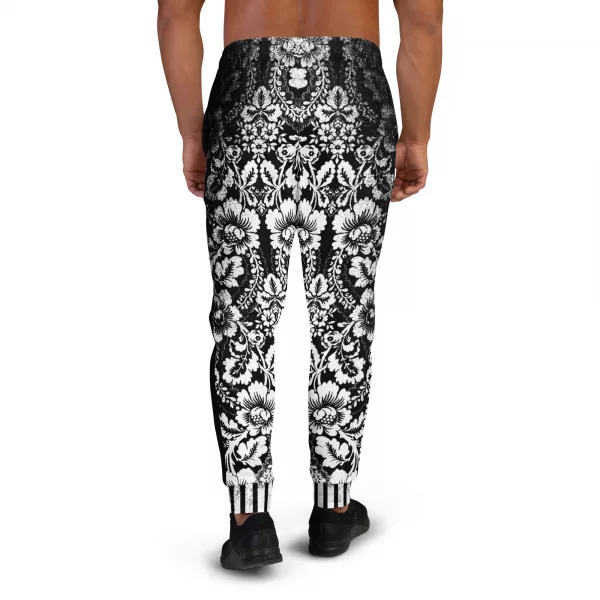 Ein Mann steht vor einem weißen Hintergrund und trägt eine schwarze Jogginghose der Marke ANTONY YORCK mit einem auffälligen weißen floralen Muster im baroque Noir Stil. Die Hose weist den Schriftzug "ANTONY YORCK" am Bein auf und hat ein gestreiftes Bündchen. Der Mann kombiniert sie mit schwarzen Sportschuhen. Perfekt für luxury urban street style, streetwear fashion, subversive style und hiphop style Liebhaber. Entdecken Sie mehr auf https://antonyyorck.com. Schlüsselwörter: trendy menswear, modern fashion, designer joggers, urban clothing, stylish activewear.