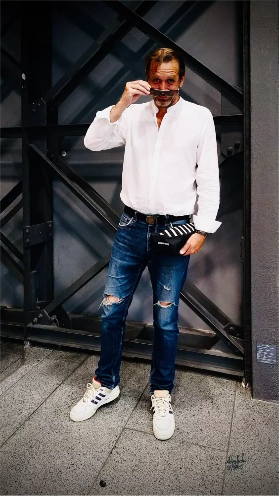 Der Designer Antony Yorck trägt ein weißes Hemd, blaue Jeans und weiße Sneakers, hält eine Bauchtasche vom eigenen Label in seiner linken Hand, nimmt mit der rechten Hand seine Sonnenbrille ab und steht vor einer Industriellen statischen Konstruktion einer Fabrikhalle.